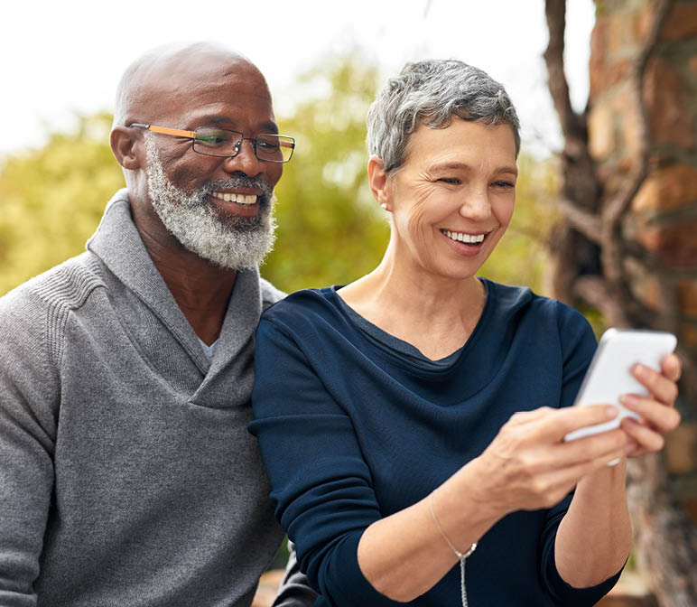 Interracial senior couple, phone and smile in park, garden or nature for love or care together. Happy man, woman and partner for retirement, relax and support for marriage and vacation for pensioner.